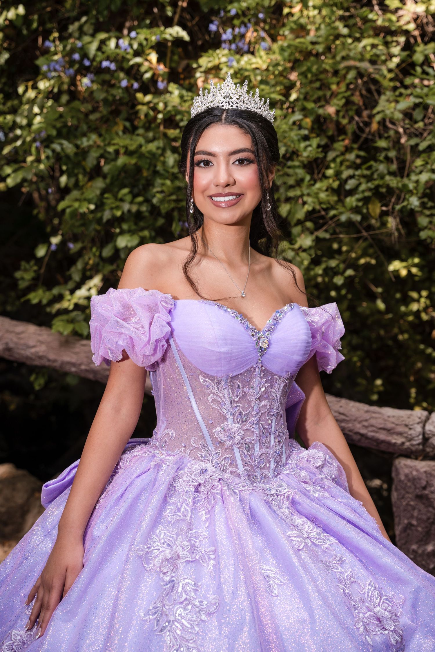 A stunning lilac quinceañera ball gown with intricate lace details and puffy sleeves photographed in a garden setting.