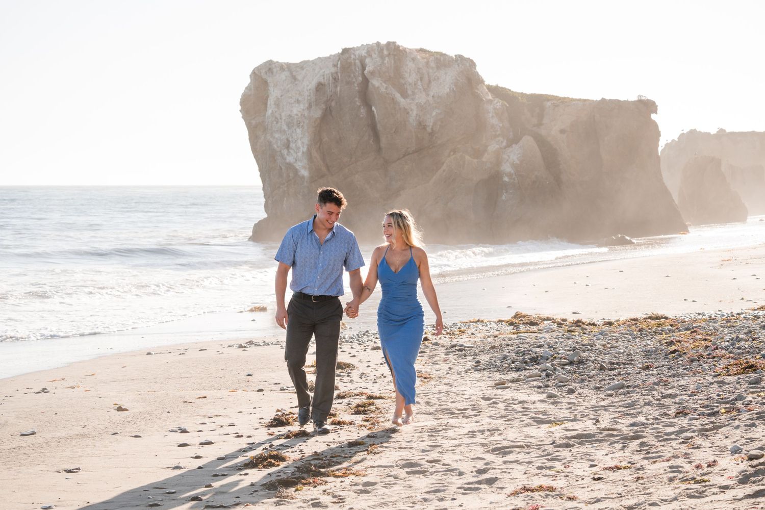 Couple walking hand in hand along a scenic beach with rock formations.