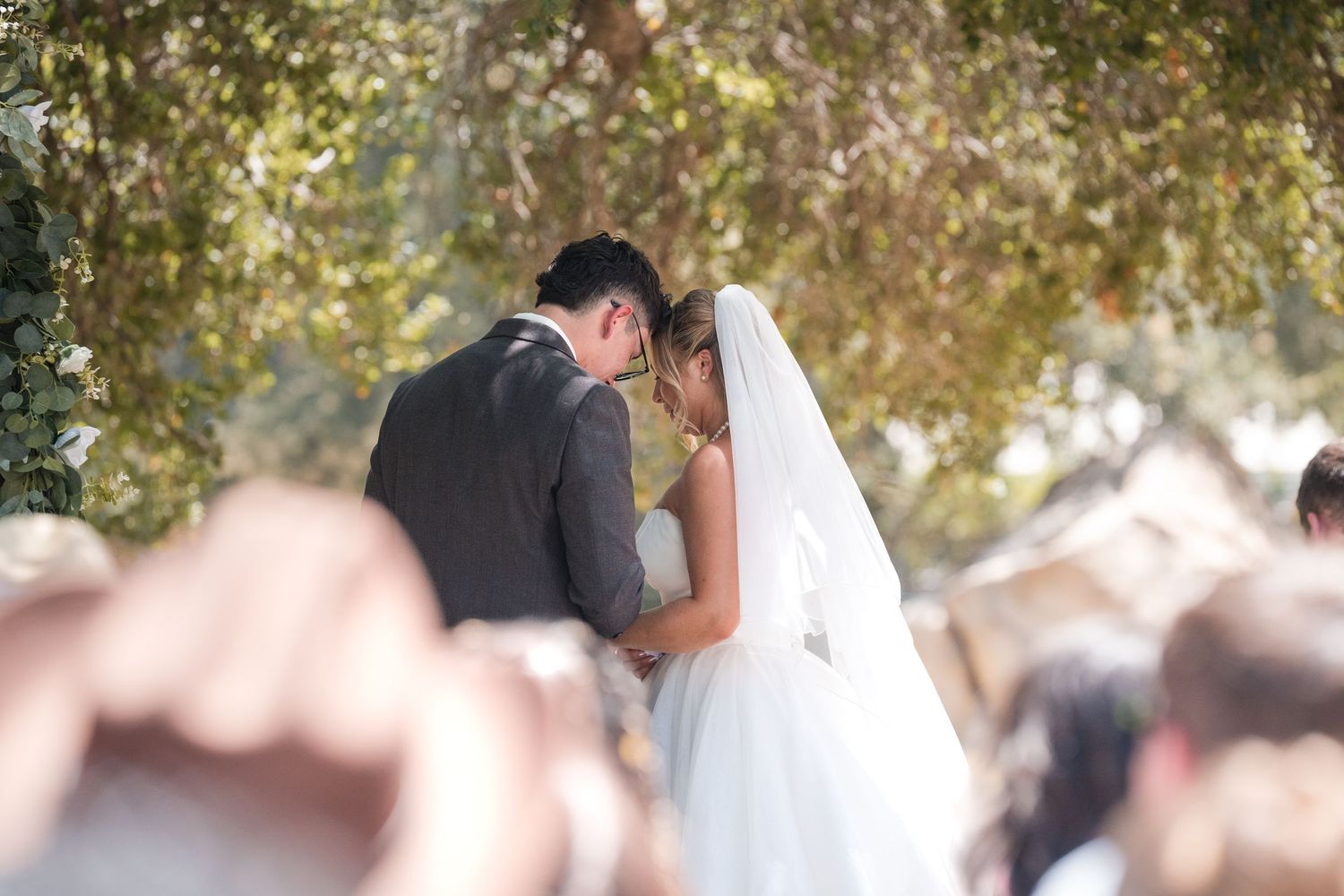Bride and groom sharing a first kiss during outdoor wedding ceremony under lush green trees.