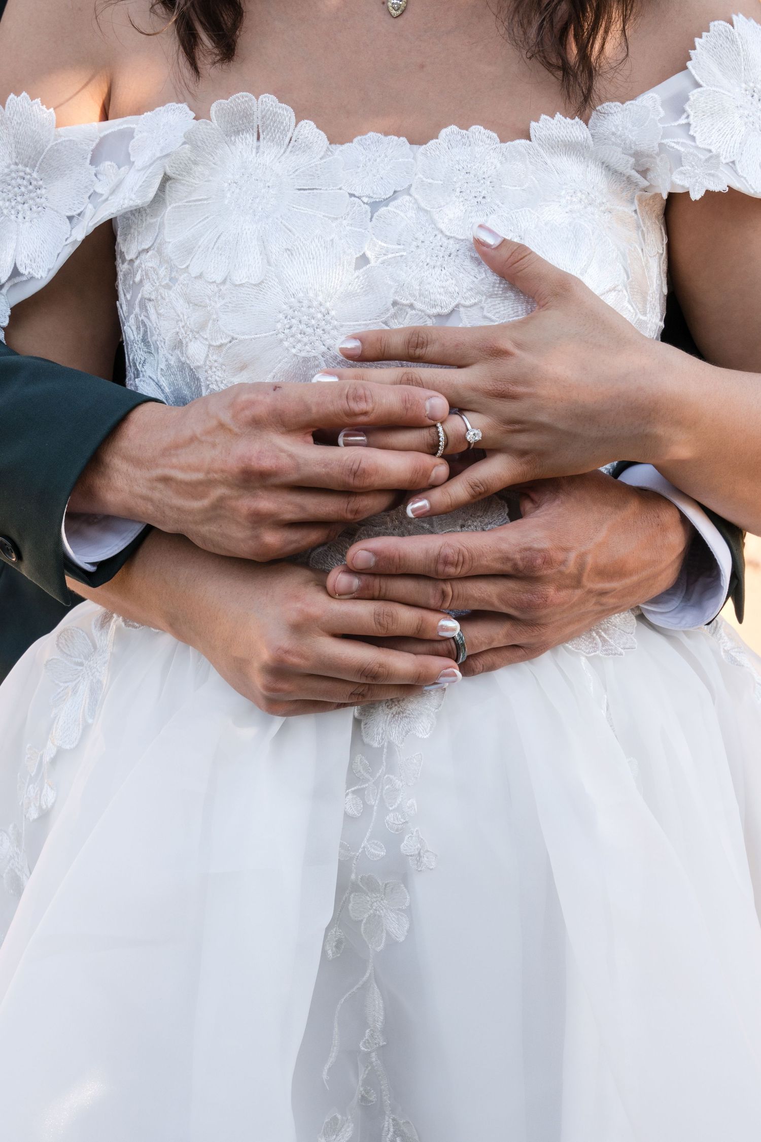 Close-up of hands with wedding rings against white lace wedding dress.