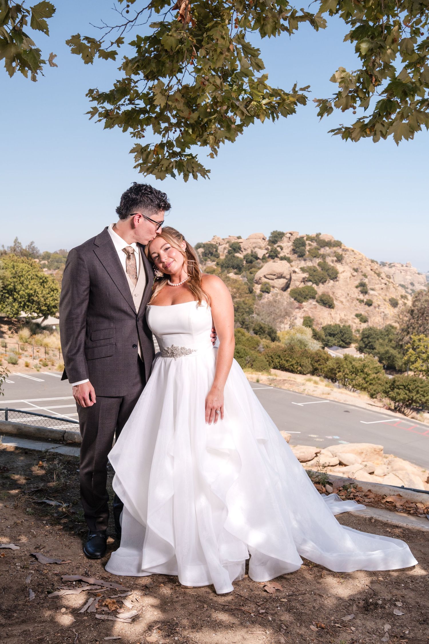 Bride in white ball gown and groom in brown suit pose romantically outdoors with scenic hills and blue sky backdrop.