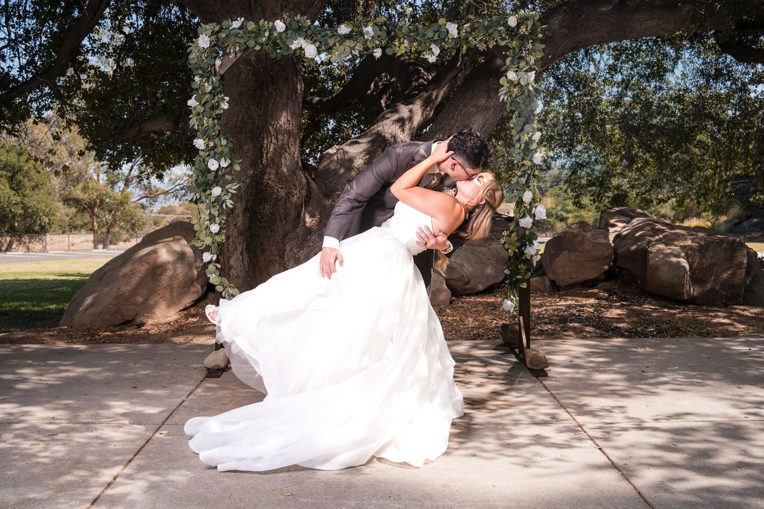 Bride and groom share a romantic dip kiss under a floral arch beside a large tree on their wedding day.