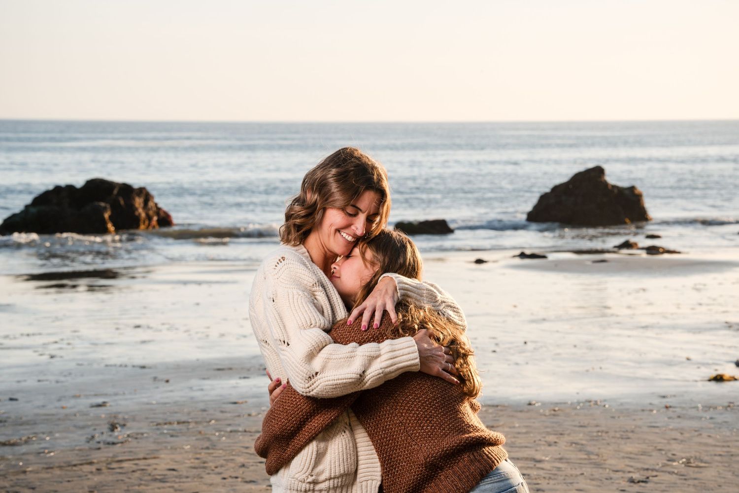 Person in cream sweater hugging and laughing on a beach at sunset with rocks in background.