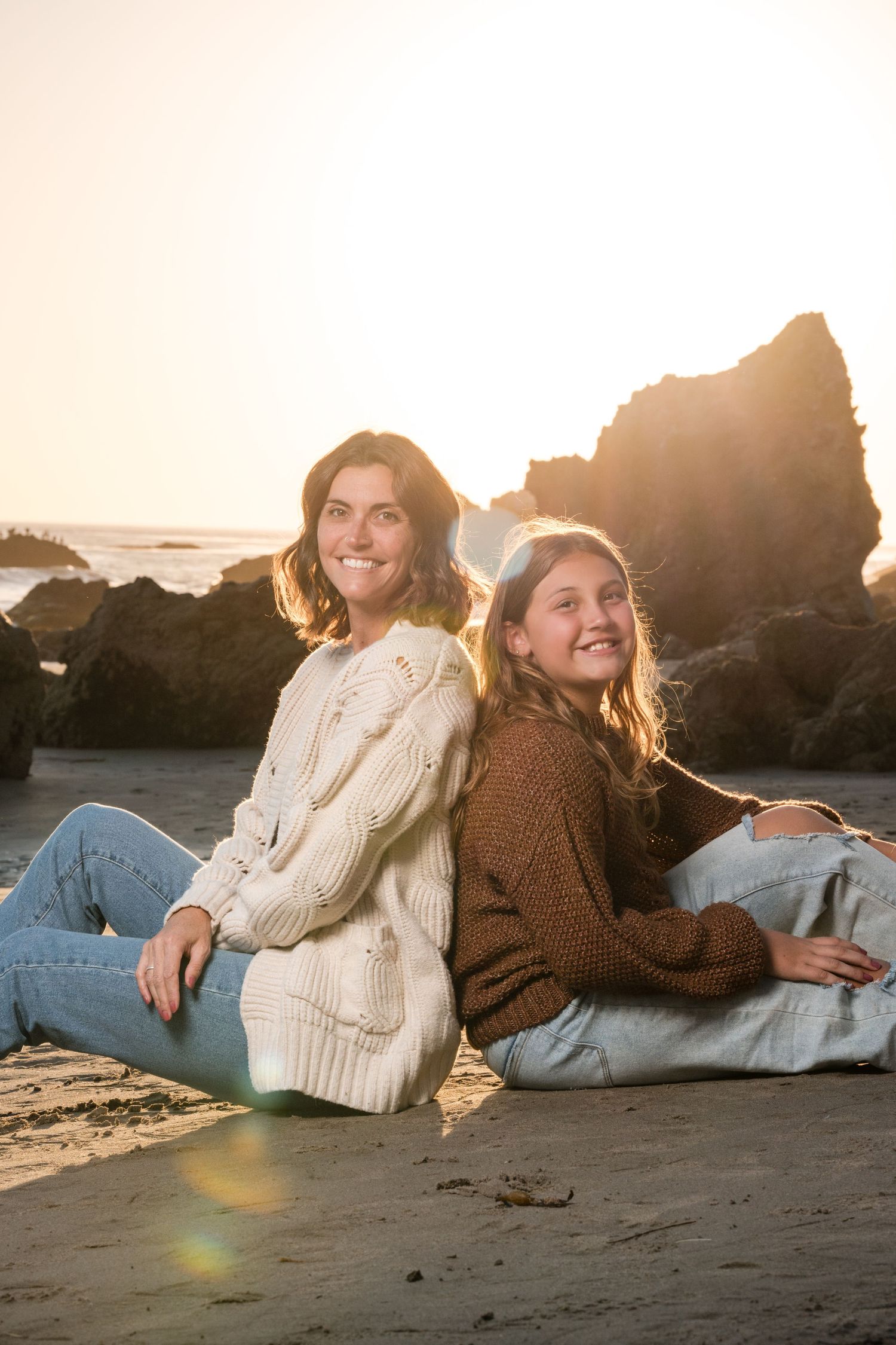 Two people in sweaters sitting back-to-back on beach sand during golden hour with rocky coastline.