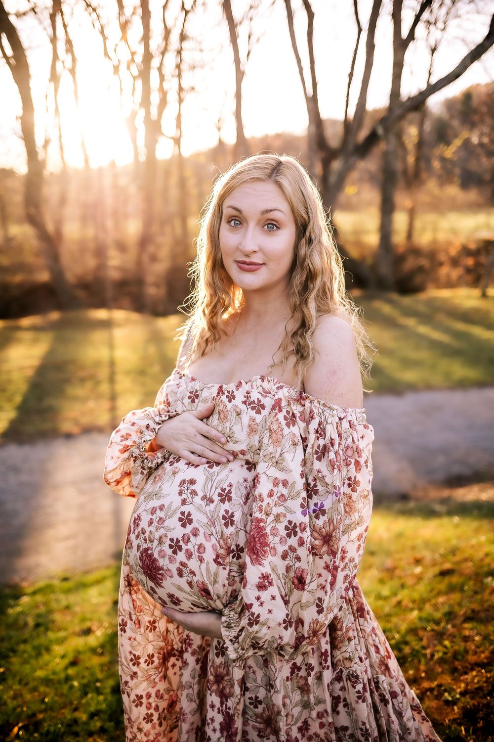 Ethereal maternity portrait of a woman in a floral off-shoulder dress standing in golden sunlit woodland setting.