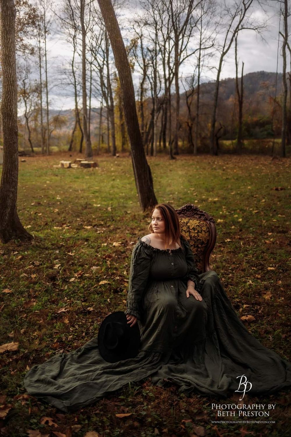 Dramatic maternity photo with vintage chair in autumn forest setting.