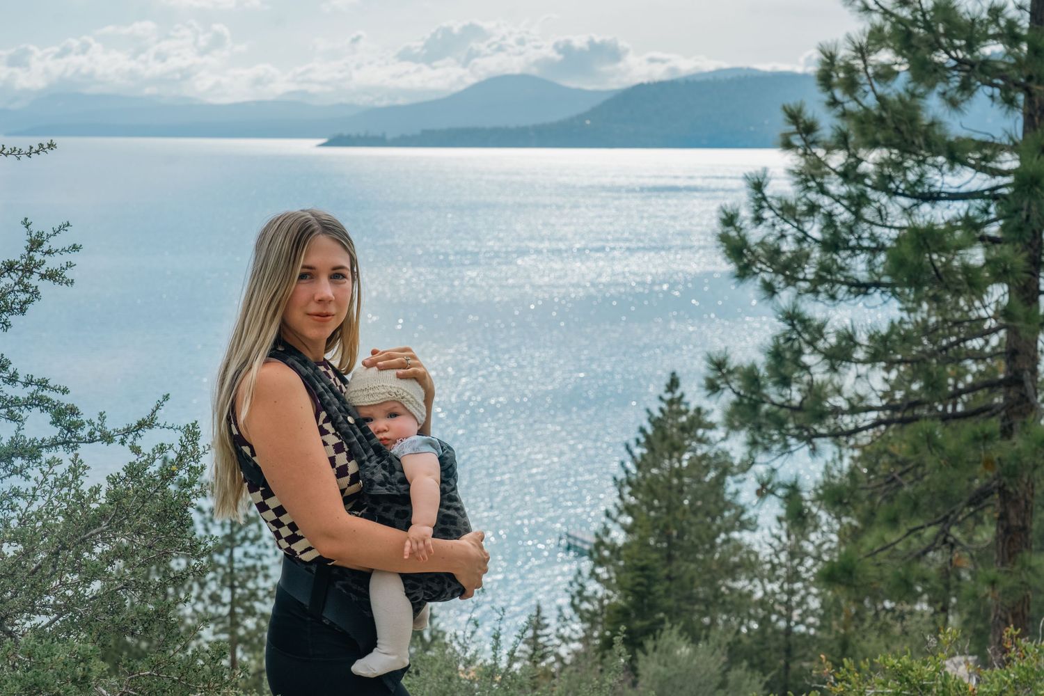 New Born child and her mother stand on a scenic mountain overlook with evergreen trees and sparkling lake tahoe in the background.