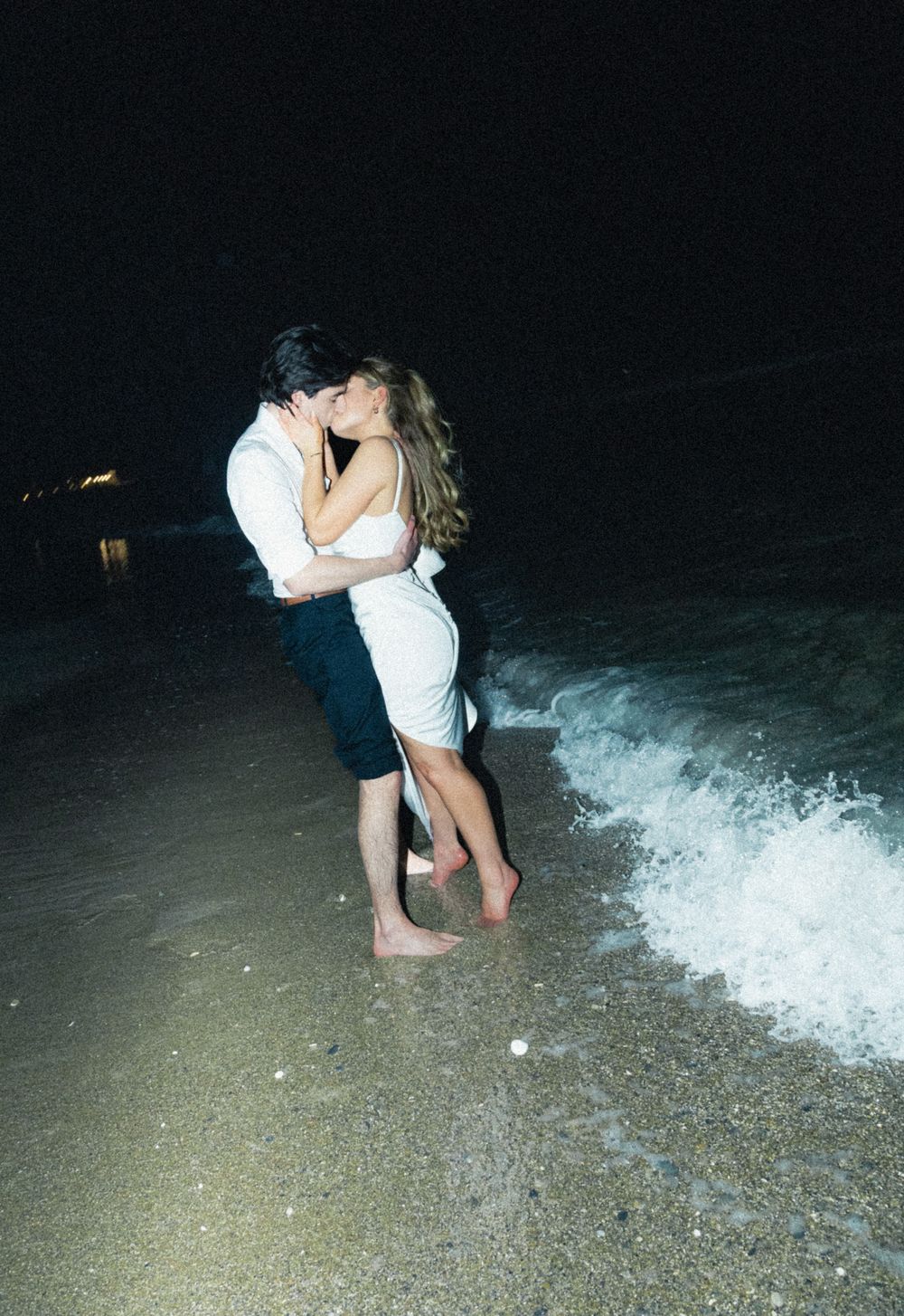 A romantic couple embraces on a beach at night as waves crash on the shore in the darkness.