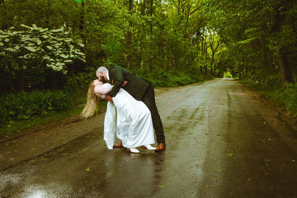 Bride and groom sharing a romantic dip kiss on a rain-soaked road surrounded by lush green trees.