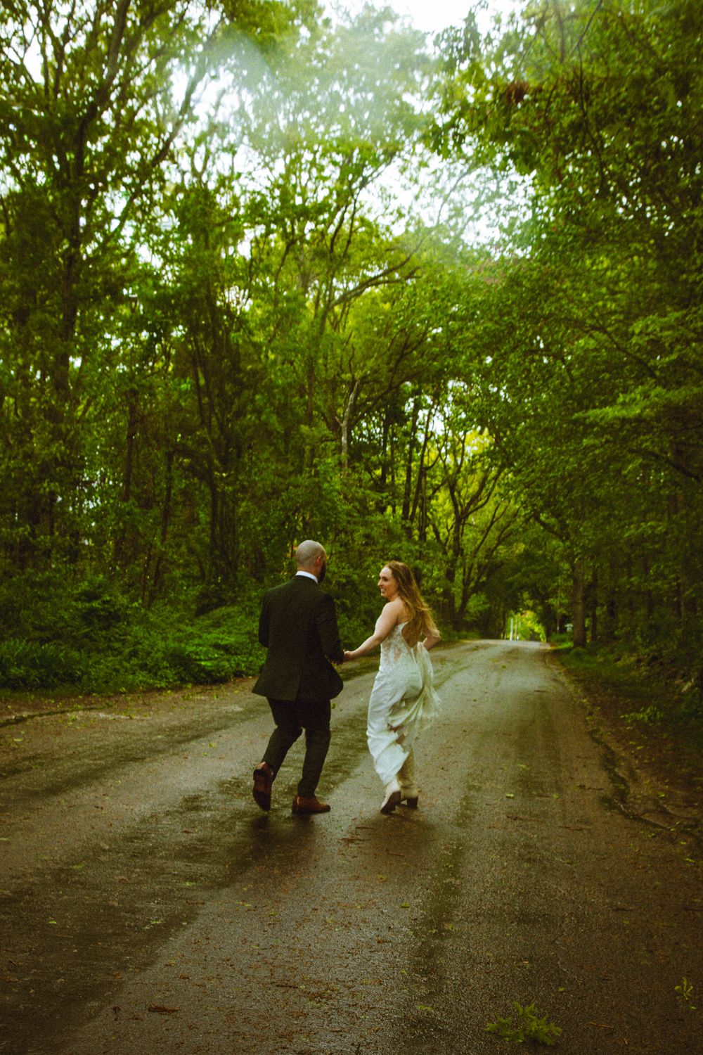Bride and groom walking hand in hand down a lush, tree-lined road on their wedding day.