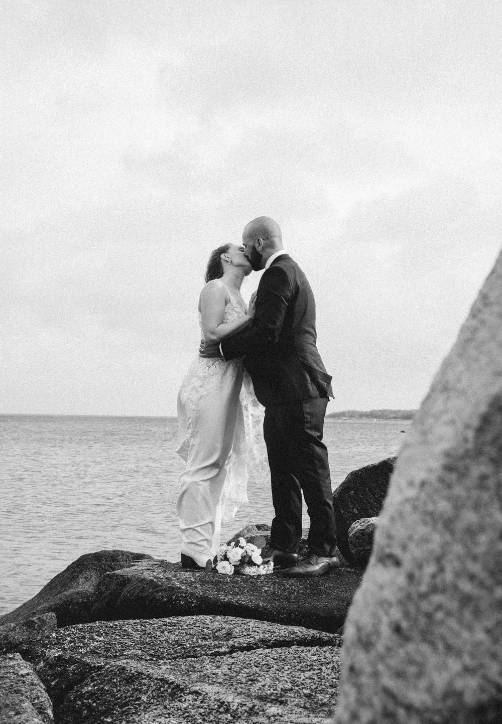 Black and white photo of a bride and groom kissing on rocky shoreline by the sea on their wedding day.