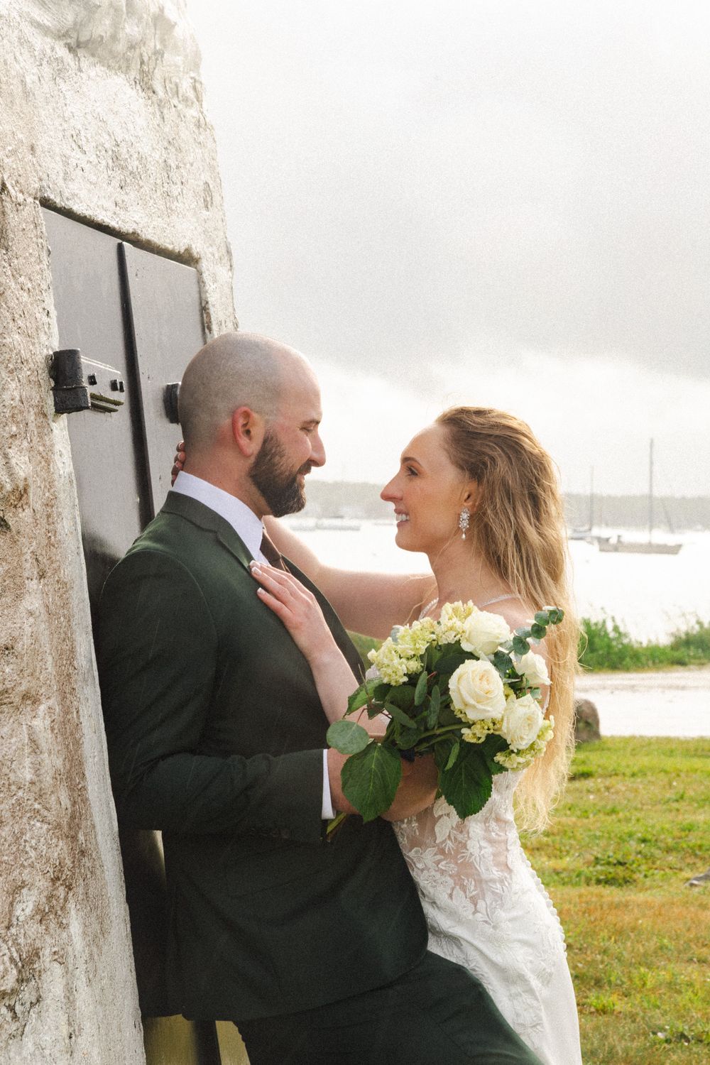 Bride and groom share a romantic moment by a stone wall near the ocean, bride holding white roses bouquet.