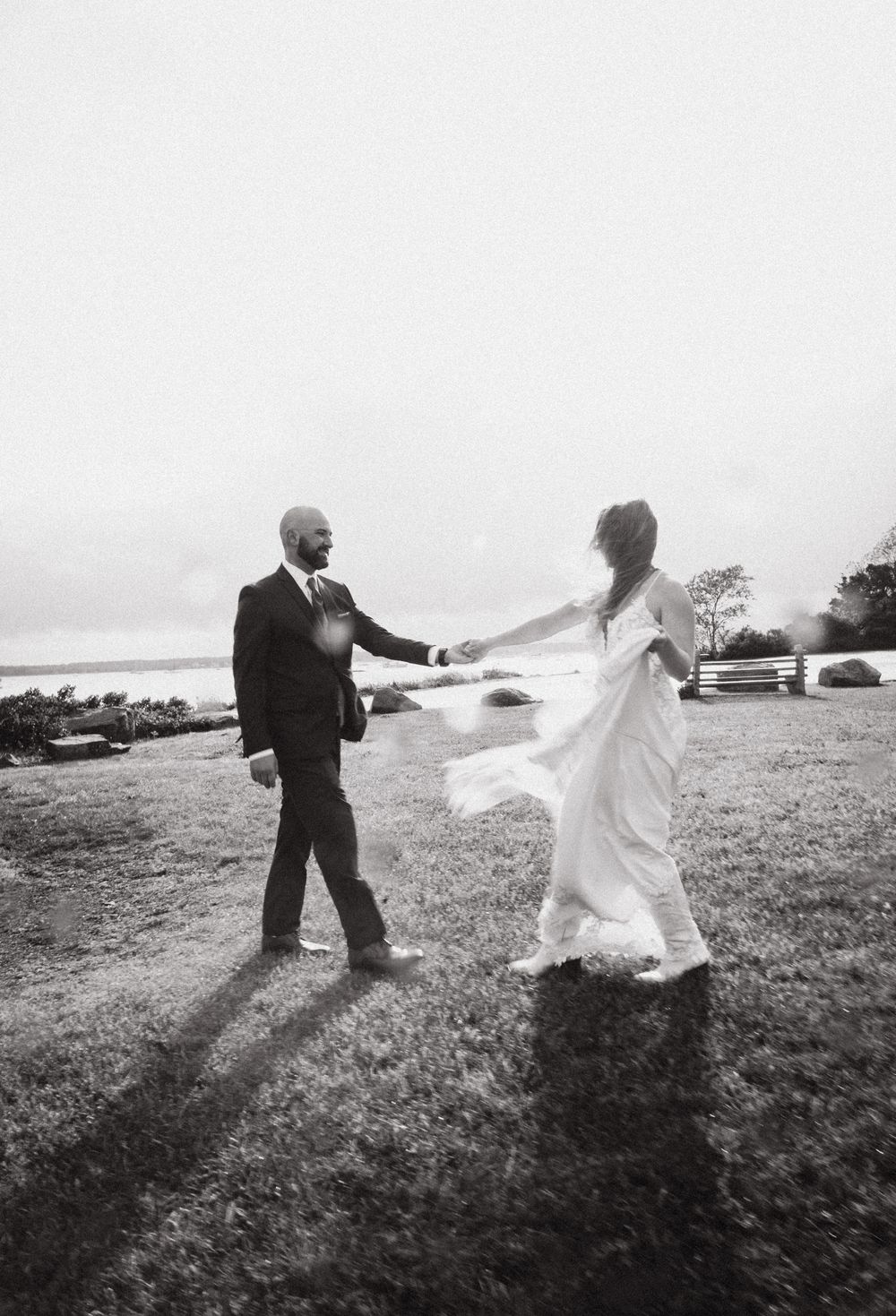 Black and white photo of a bride and groom dancing outdoors on grass near the water on their wedding day.