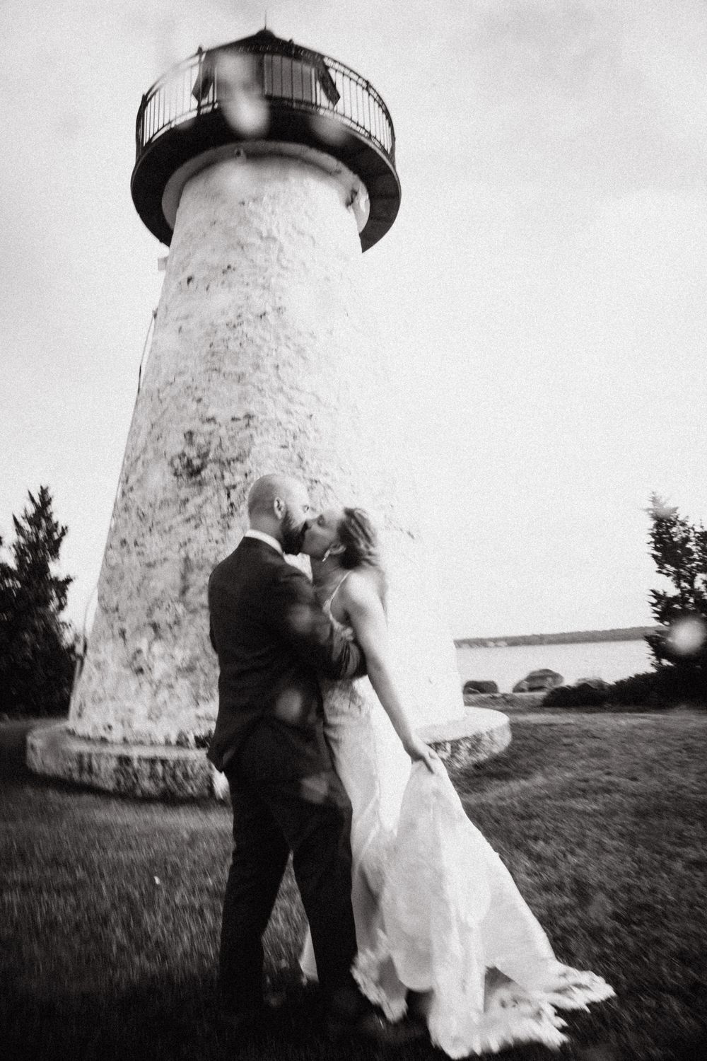 Black and white photo of a bride and groom kissing in front of a historic lighthouse on their wedding day.