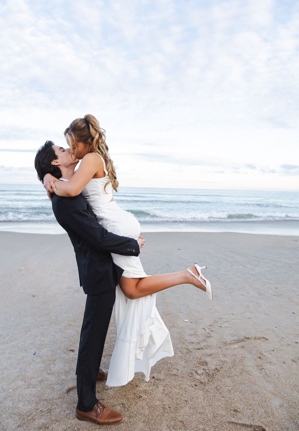 A couple in wedding attire embraces and kisses on a scenic beach with ocean waves in the background.