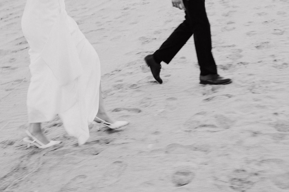 Couple walking on a beach in formal wedding attire with their feet in the sand and water.