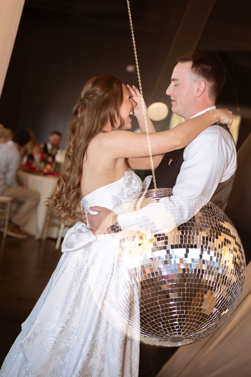 A couple dances beneath a sparkling disco ball at a wedding reception, with the bride wearing a flowing white dress.