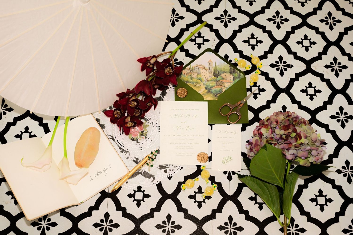 Wedding stationery flat lay with green envelope, floral invitation, white parasol, and hydrangeas on black and white patterned surface.