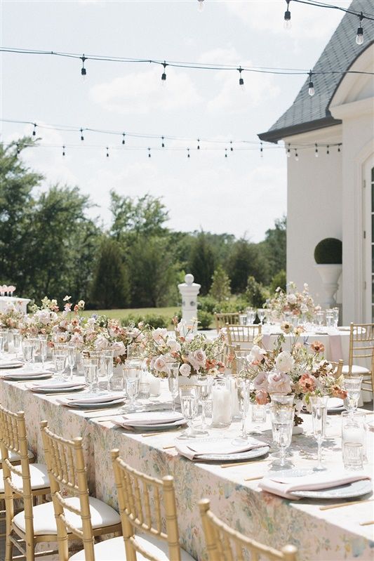 Elegant outdoor wedding reception table with string lights, gold chairs, white linens and soft pink floral centerpieces.