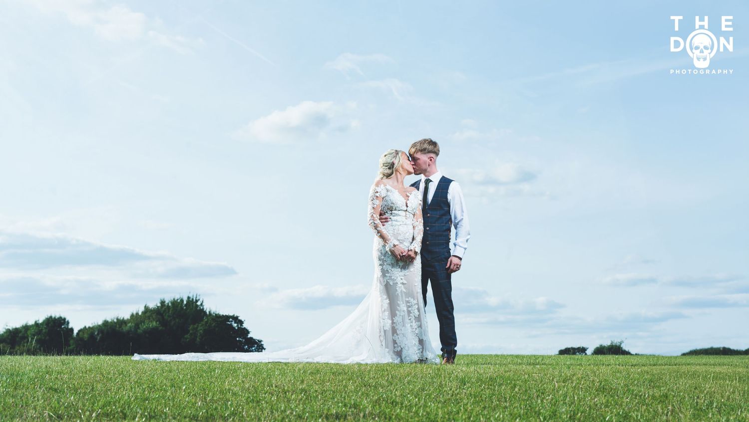 Bride and groom standing on lush green grass under blue sky at outdoor wedding venue, captured by The Don.