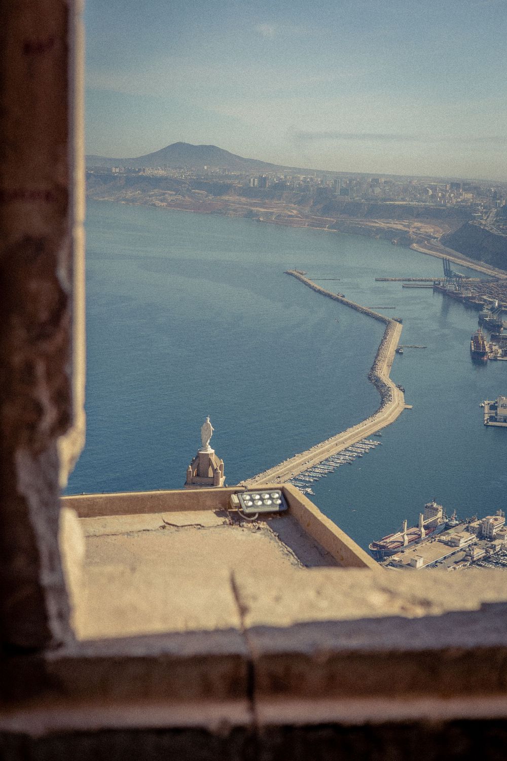 Harbor breakwater and port facilities viewed through a window frame with mountains in the distance.