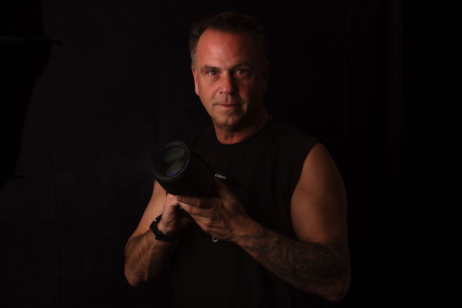 A muscular man in a sleeveless shirt holds a camera in a dramatic dark studio setting.