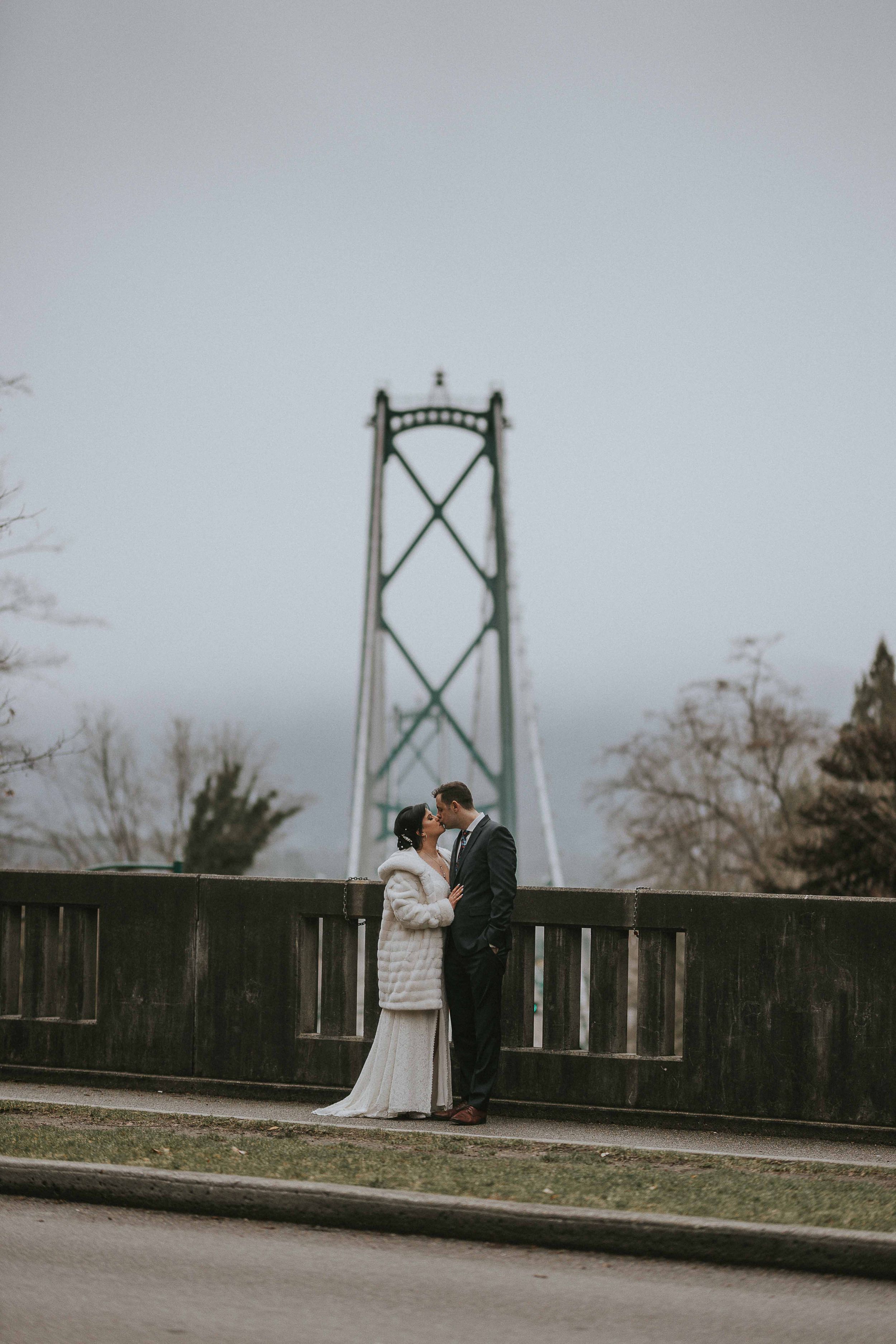 Couple shares romantic moment on bridge with iconic Lions Gate Bridge tower rising in misty background.