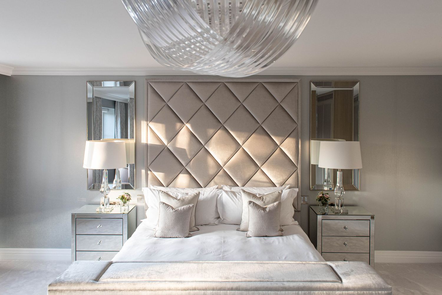 A luxurious bedroom interior with mirrored panels, crystal chandelier, and elegant taupe upholstered headboard.
