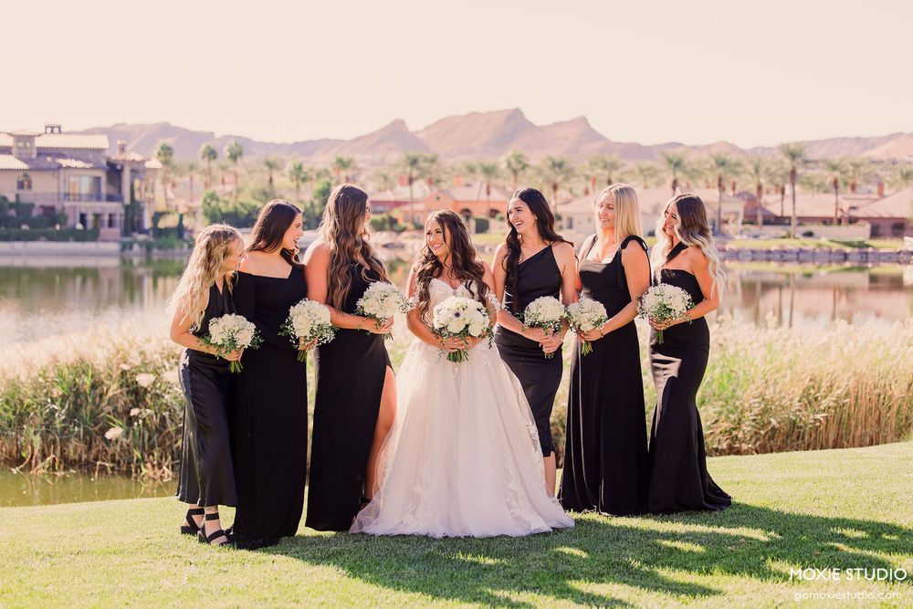Wedding party members in black dresses and formal attire pose together on a scenic grassy overlook with mountains in background.