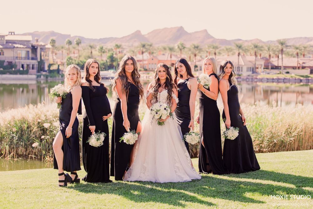Wedding party in black bridesmaid dresses poses with bride in white gown on golf course with lake and mountains in background.