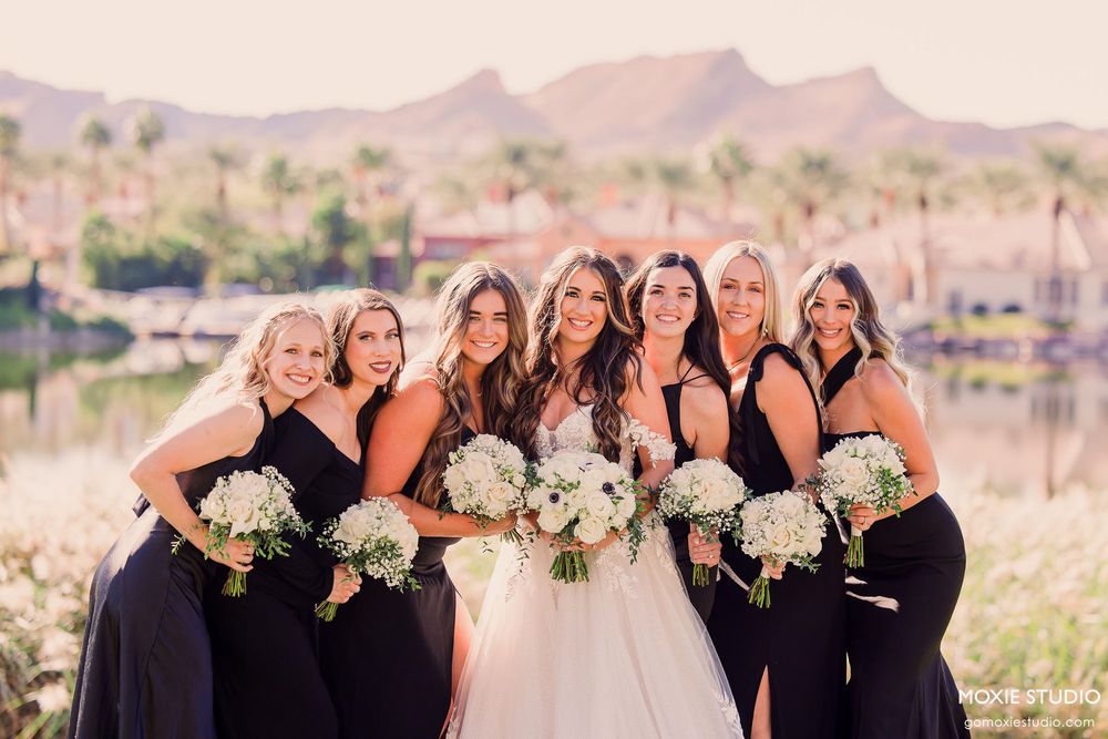 A group of bridesmaids in black dresses pose with bouquets against a desert mountain backdrop at sunset.