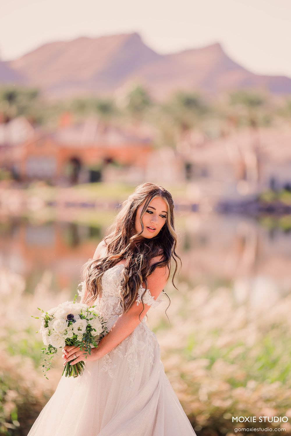 A bride in a white lace wedding dress holds a bouquet of white flowers against a desert mountain backdrop at sunset.