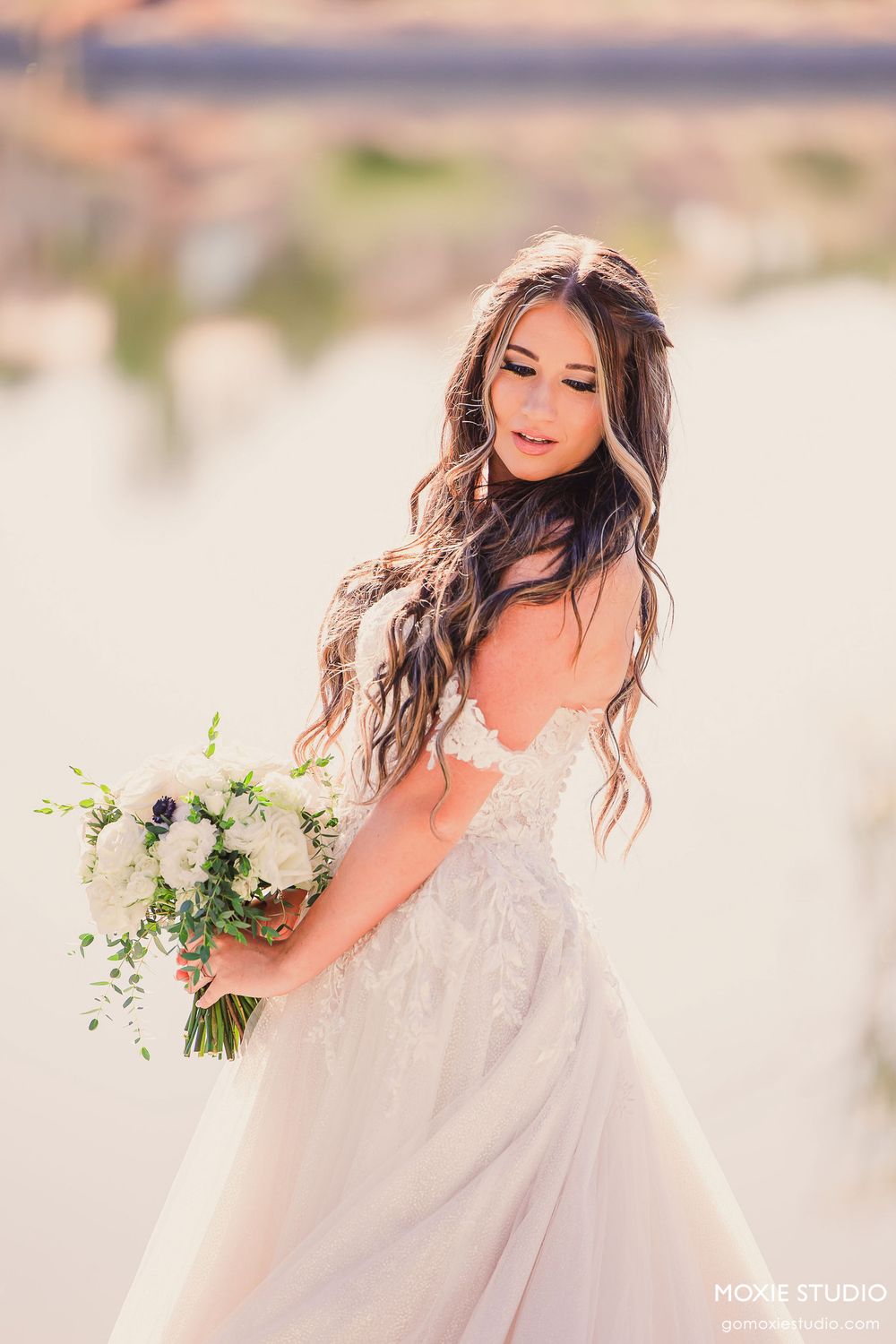 A bride in a flowing white wedding gown poses with a bouquet against a dreamy beach backdrop.