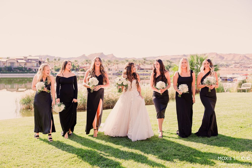 Wedding party in black dresses and a bride in white pose together on a grassy lawn with mountains in the background.