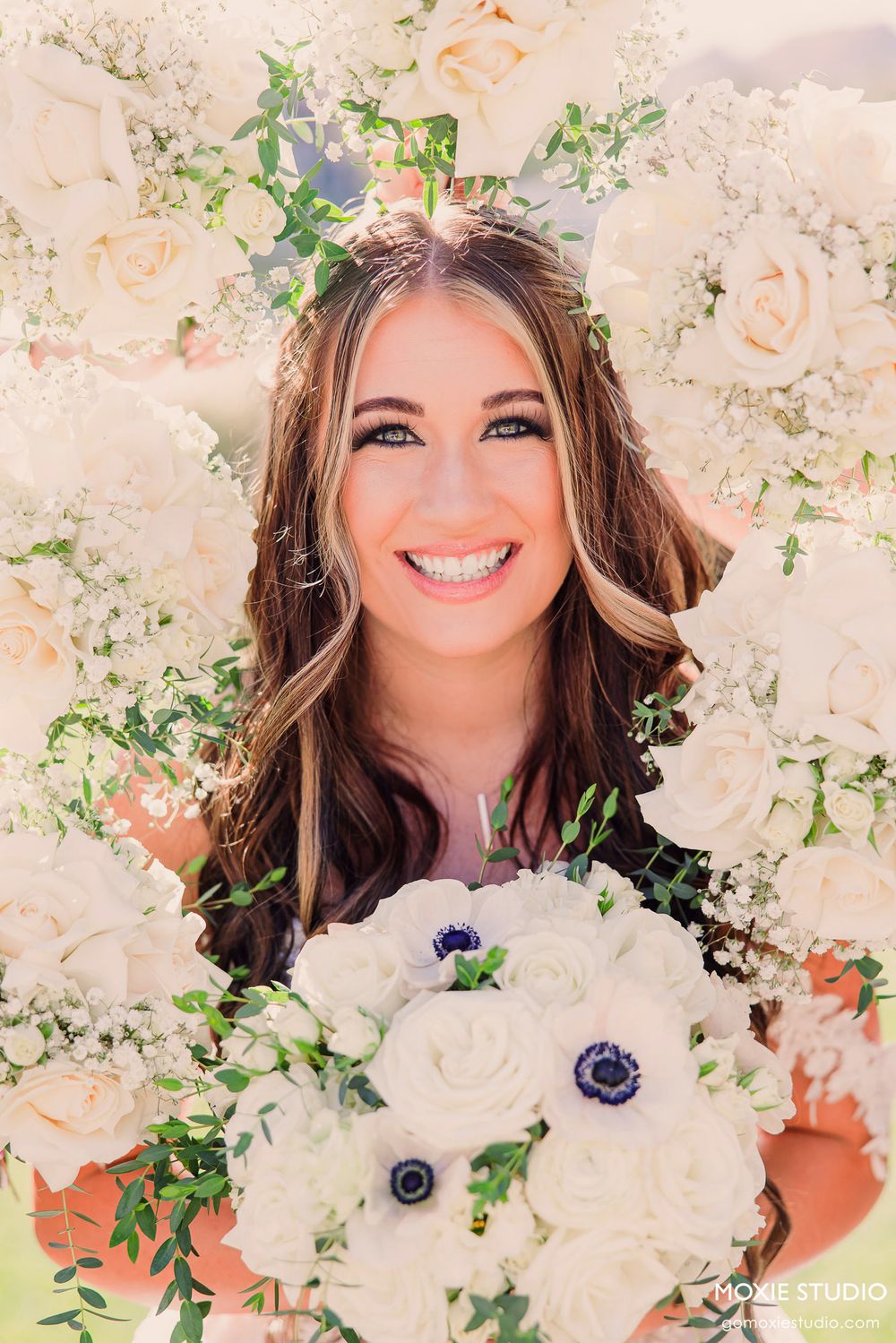 Bridal portrait with white roses and anemone bouquet surrounded by floral backdrop and delicate greenery crown.