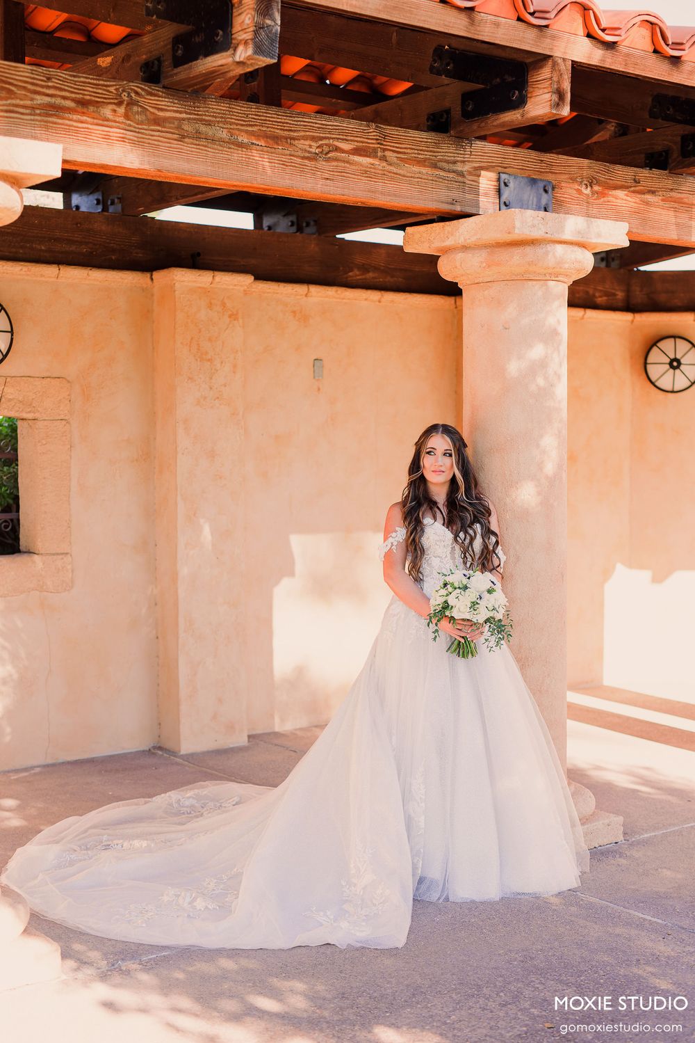 A bride in a flowing white wedding gown stands under a rustic covered patio with exposed wooden beams and peach-colored walls.