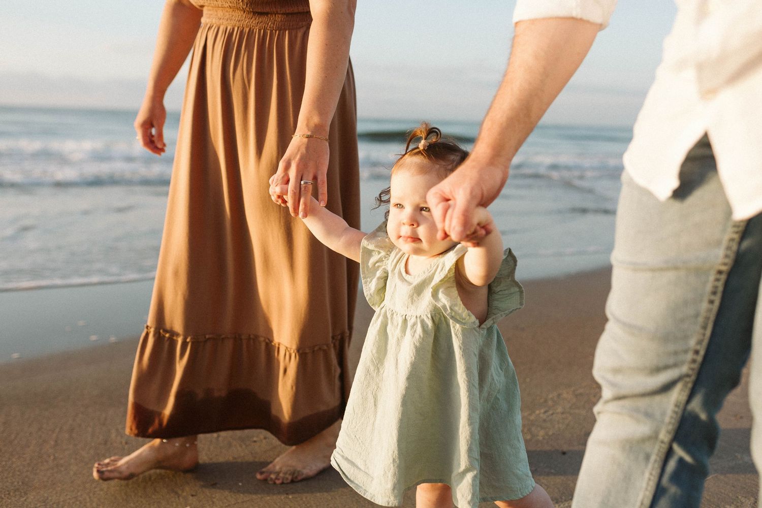 Photo by B. Logsdon Photography of a small child in a light dress holds hands with parents while walking on a sandy beach at sunset.