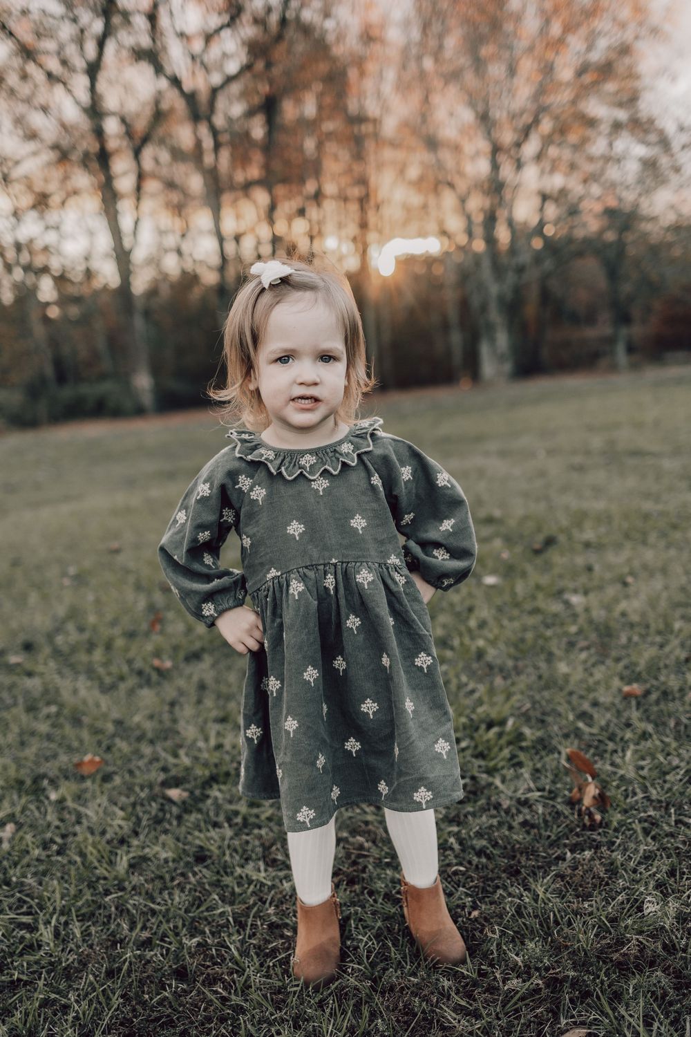 A toddler in a green floral dress and brown boots stands in a grassy field during autumn sunset.