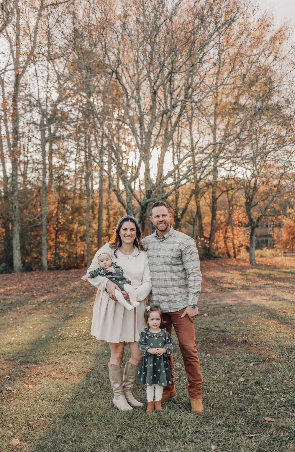 Family poses together in autumn setting wearing coordinated neutral outfits against backdrop of fall foliage.