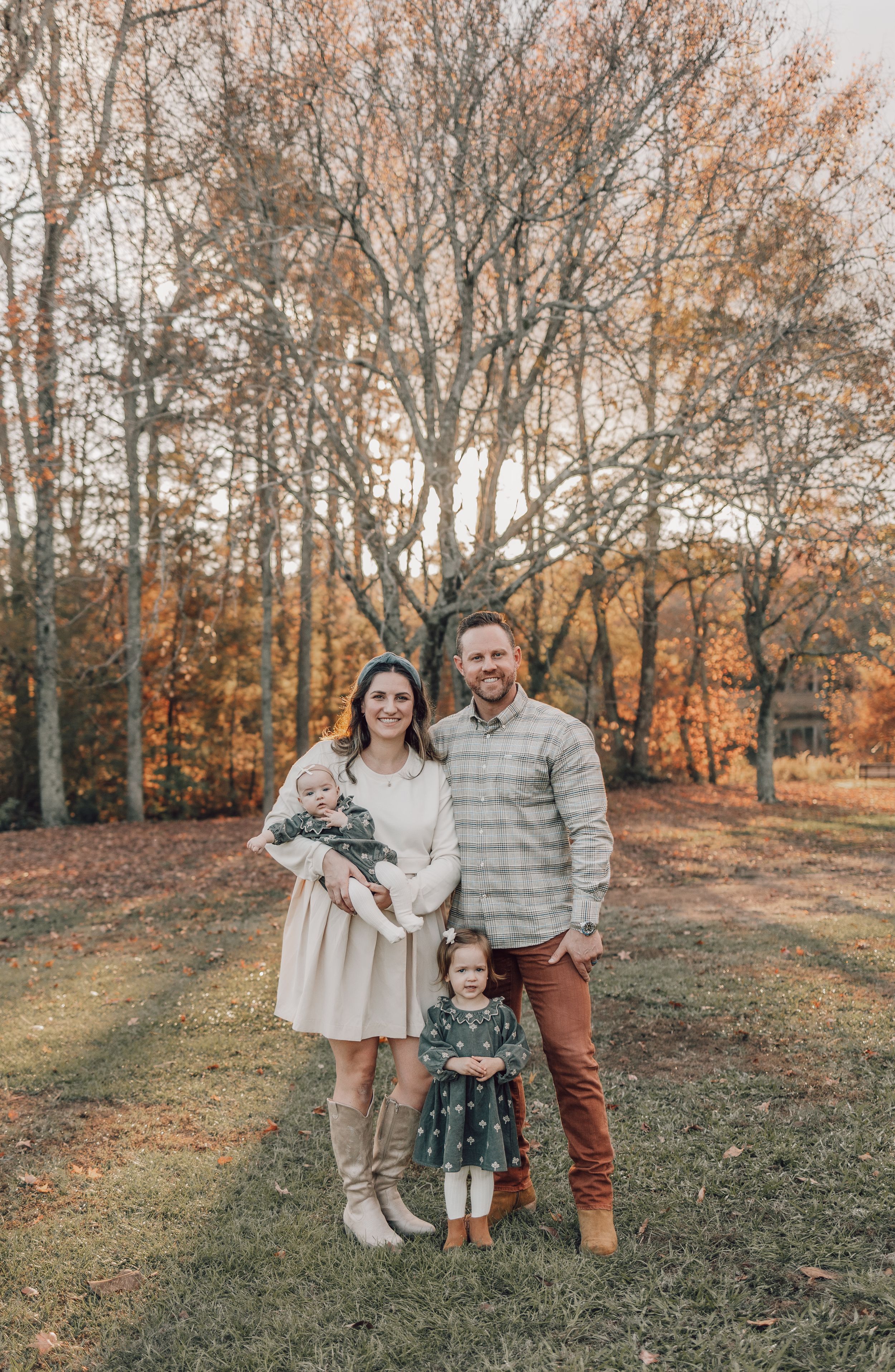 Family poses together in autumn setting wearing coordinated neutral outfits against backdrop of fall foliage.