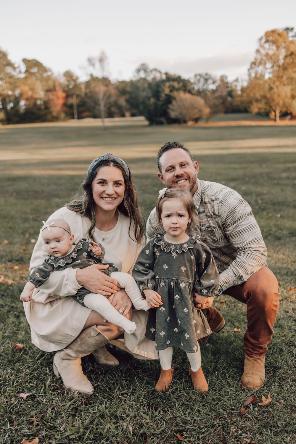 Family sits together on grassy field during golden hour photoshoot in coordinated neutral outfits.