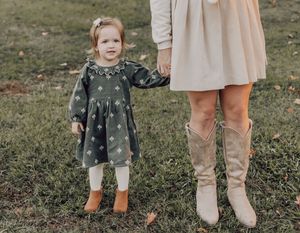 A child in a polka dot dress stands next to someone wearing a beige dress and tall boots on grass.