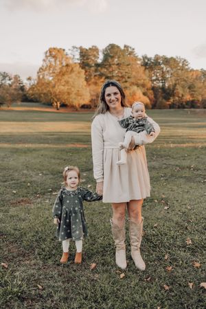 Family members pose together on grass during a fall photoshoot with trees displaying autumn colors.
