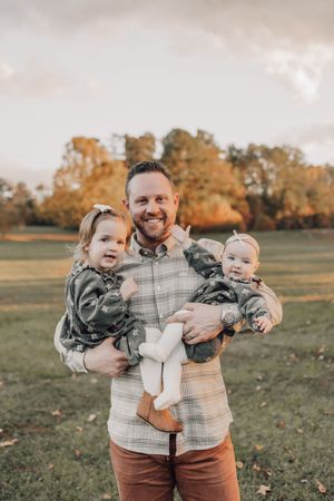 Family portrait in autumn field with parent holding two small children wearing coordinating outfits.