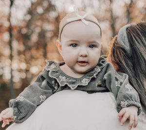 Sweet baby wearing a ruffled camo dress and headband smiles while being held against autumn trees in the background.