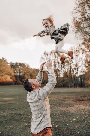 Parent playfully lifts child in air during autumn outdoor photoshoot.