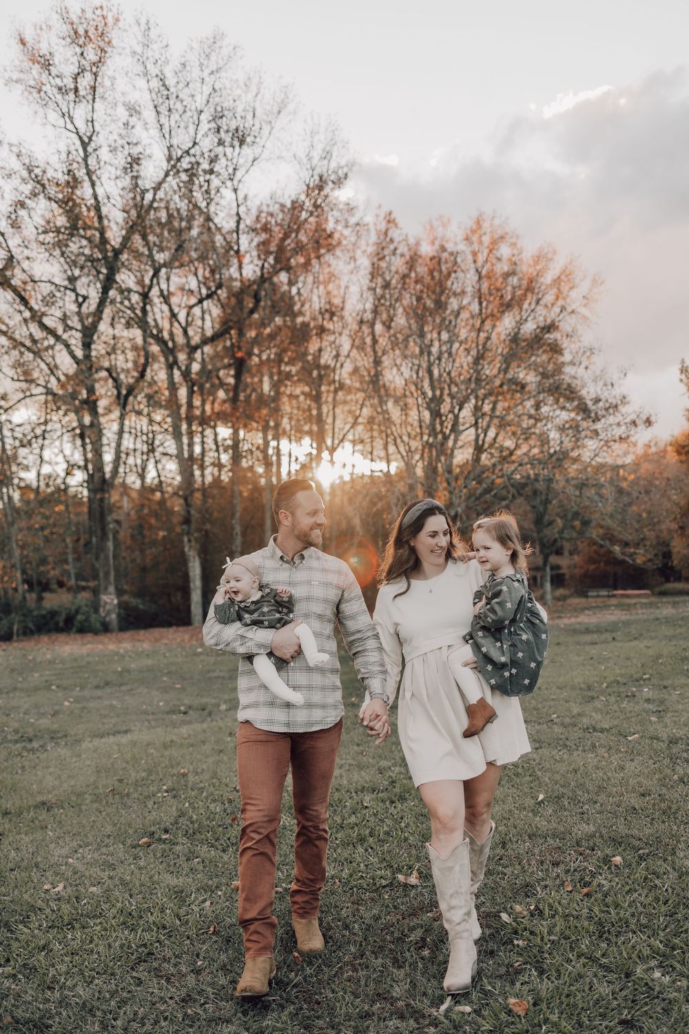 A couple walks hand in hand through an autumn field at sunset with bare trees in the background.