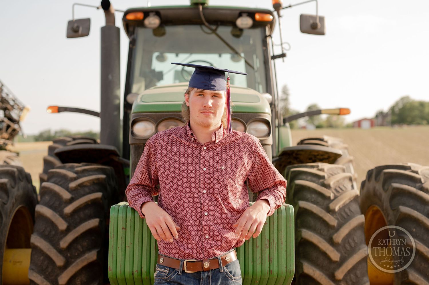 Person in baseball cap and red shirt stands in front of large green tractor.