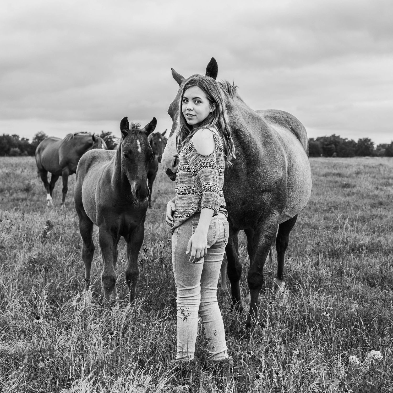 Young person in denim stands in a field with two brown horses on an overcast day.