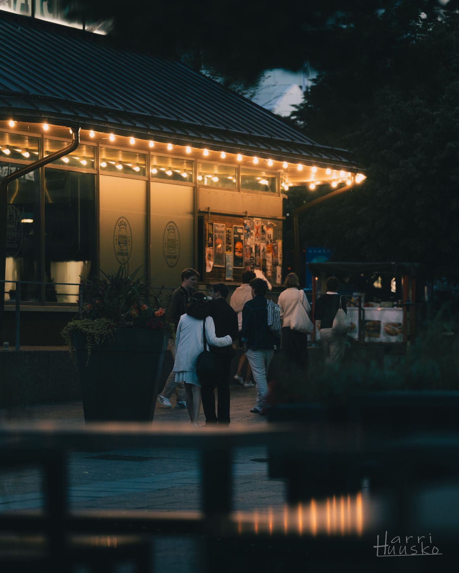 Evening gathering under string lights at a cozy outdoor venue with reflections in water.